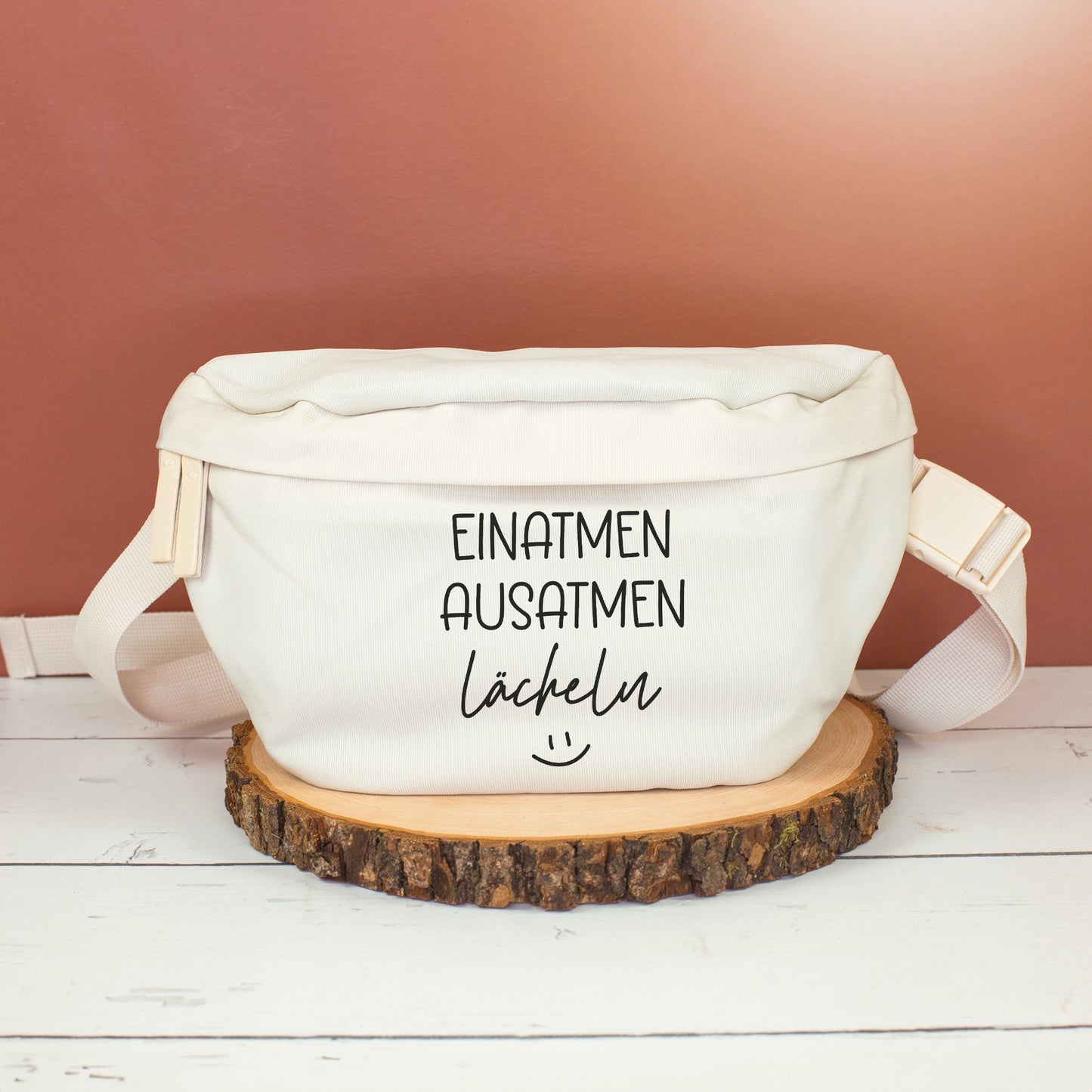 Cremefarbene Bauchtasche mit Spruch Einatmen Ausatmen Lächeln frontal auf Holzscheibe vor rötlicher Wand