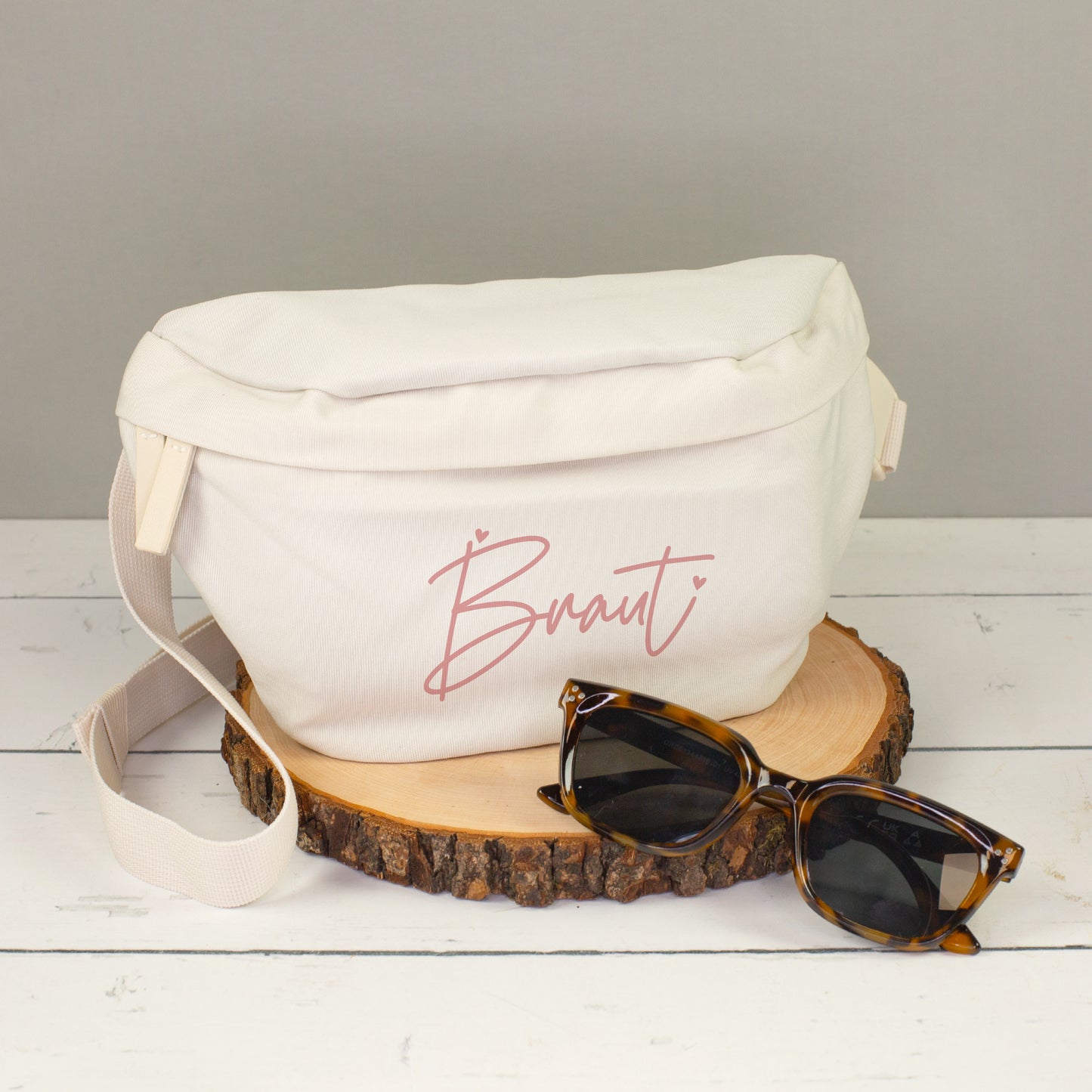 Cremefarbene Bauchtasche mit rosa Braut-Aufdruck auf Holzscheibe, dekoriert mit Sonnenbrille
