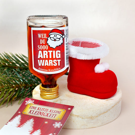 Detailaufnahme eines roten Nikolausstiefels mit weißem Kunstfellrand und Pompons. Daneben steht eine auf dem Kopf platzierte Likörflasche mit dem Spruch „Weil du sooo artig warst“. Im Vordergrund eine Karte mit Weihnachtsdesign.