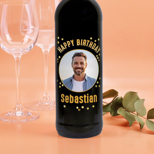 Weinflasche mit schwarzem Etikett „Happy Birthday Sebastian“ und Foto eines Mannes, dekoriert mit goldenen Punkten, vor Weingläsern.