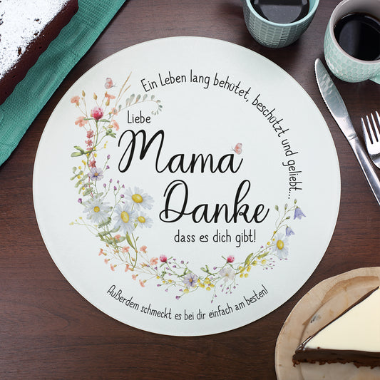 Glasbrettchen mit floraler Verzierung aus Wiesenblumen und geschwungener Schrift auf einem dunklen Holztisch, gedeckt mit Kaffeetassen und Kuchen. Der Spruch „Ein Leben lang behütet, beschützt und geliebt…“ bildet gemeinsam mit den Blumen einen harmonischen Kreis. In der Mitte steht „Liebe Mama, Danke, dass es dich gibt!“. Die personalisierte Textzeile lautet „Außerdem schmeckt es bei dir einfach am besten!“.