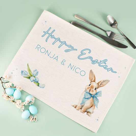 Personalisiertes Jute Platzdeckchen zu Ostern mit Hasenmotiv, blauer Schleife, Blumenillustration und individuellem Wunschtext auf mintgrünem Hintergrund