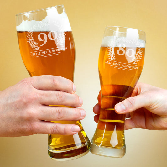Zwei Personen stoßen mit gefüllten Weizengläsern an. Beide Gläser tragen Gravuren mit Lorbeerkranz, Alter und Namen, darunter der Schriftzug „Herzlichen Glückwunsch“.