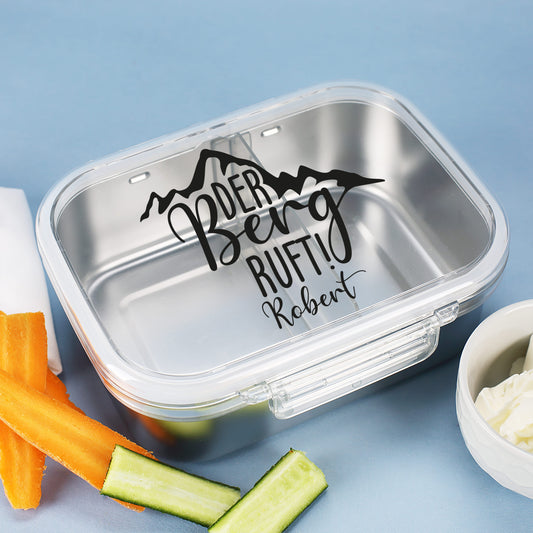 Geöffnete Brotdose mit Name und Aufdruck „Der Berg ruft!“ auf Holzuntergrund, befüllt mit Snacks