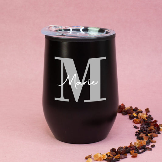 Schwarzer Thermobecher mit Initiale M und Name Marie, auf rosa Hintergrund mit Teemischung dekoriert