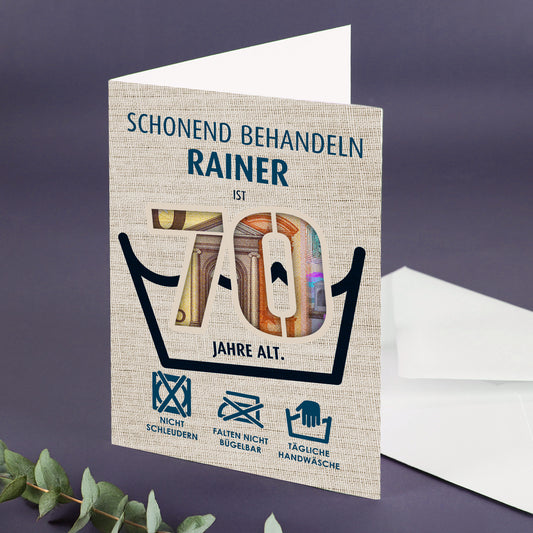 Geldgeschenk-Klappkarte mit Zahl 70 und dem Namen Rainer, aufgeklappt auf lila Untergrund neben weißem Umschlag und Eukalyptuszweig