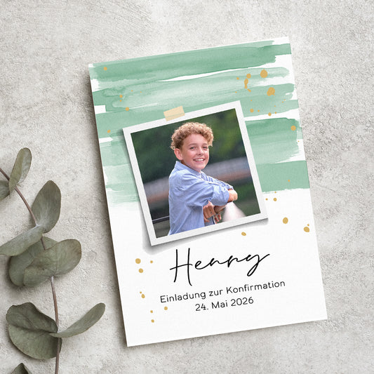 Personalisierte Einladungskarte zur Konfirmation mit modernem Aquarell-Design in Grün und Gold. Ein Polaroid-Foto des Konfirmanden ist mittig platziert, darunter sein Name und das Datum der Feier. Die Karte liegt auf einer strukturierten Steinoberfläche, umgeben von Eukalyptusblättern.
