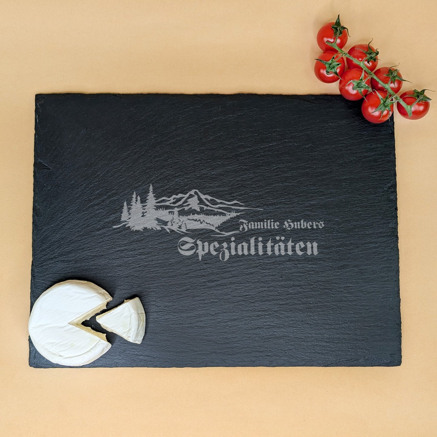 Personalisierte Schieferplatte mit Gravur „Familie Hubers Spezialitäten“ und Käse sowie Tomaten auf beigem Untergrund