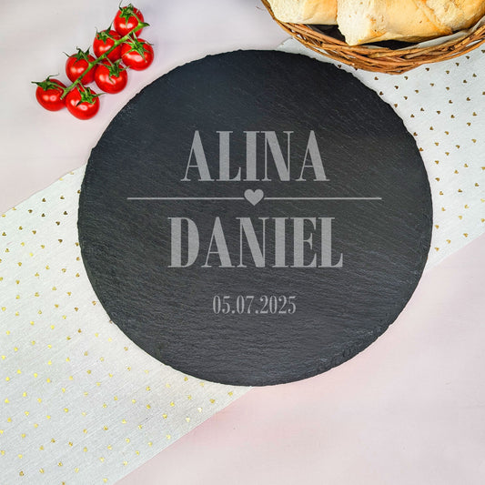 Runde Schieferplatte mit Herzmotiv Gravur, zwei Namen und Datum, präsentiert mit Baguette und Tomaten auf einem Tisch.