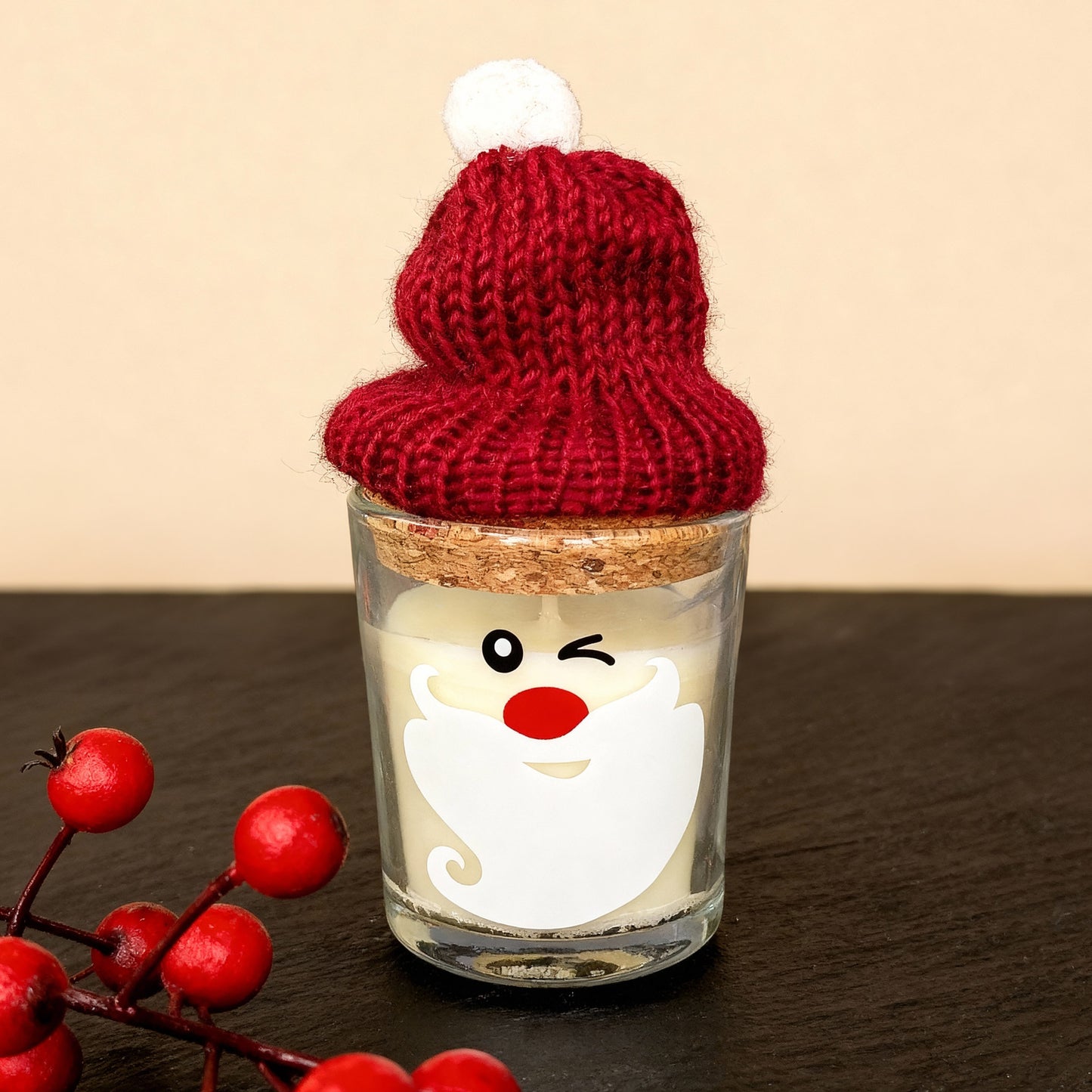 Weihnachtskerze im Glas mit fröhlichem Gesicht, roter Nase, weißem Bart und roter Pudelmütze mit Bommel – dekorative Kerze vor dunklem Hintergrund.