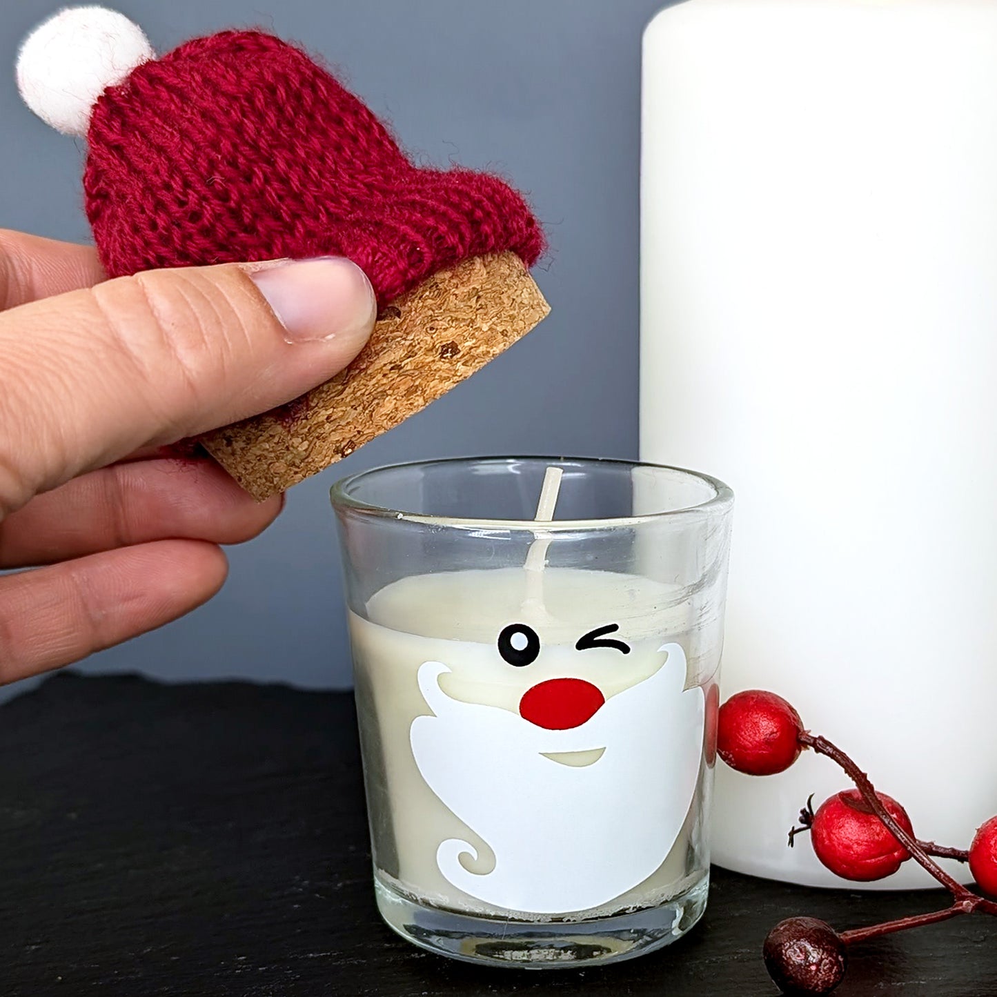 Hand hebt rote Pudelmütze mit Korkverschluss von der Kerze im Glas mit Weihnachtsgesicht – liebevolle Detailaufnahme.