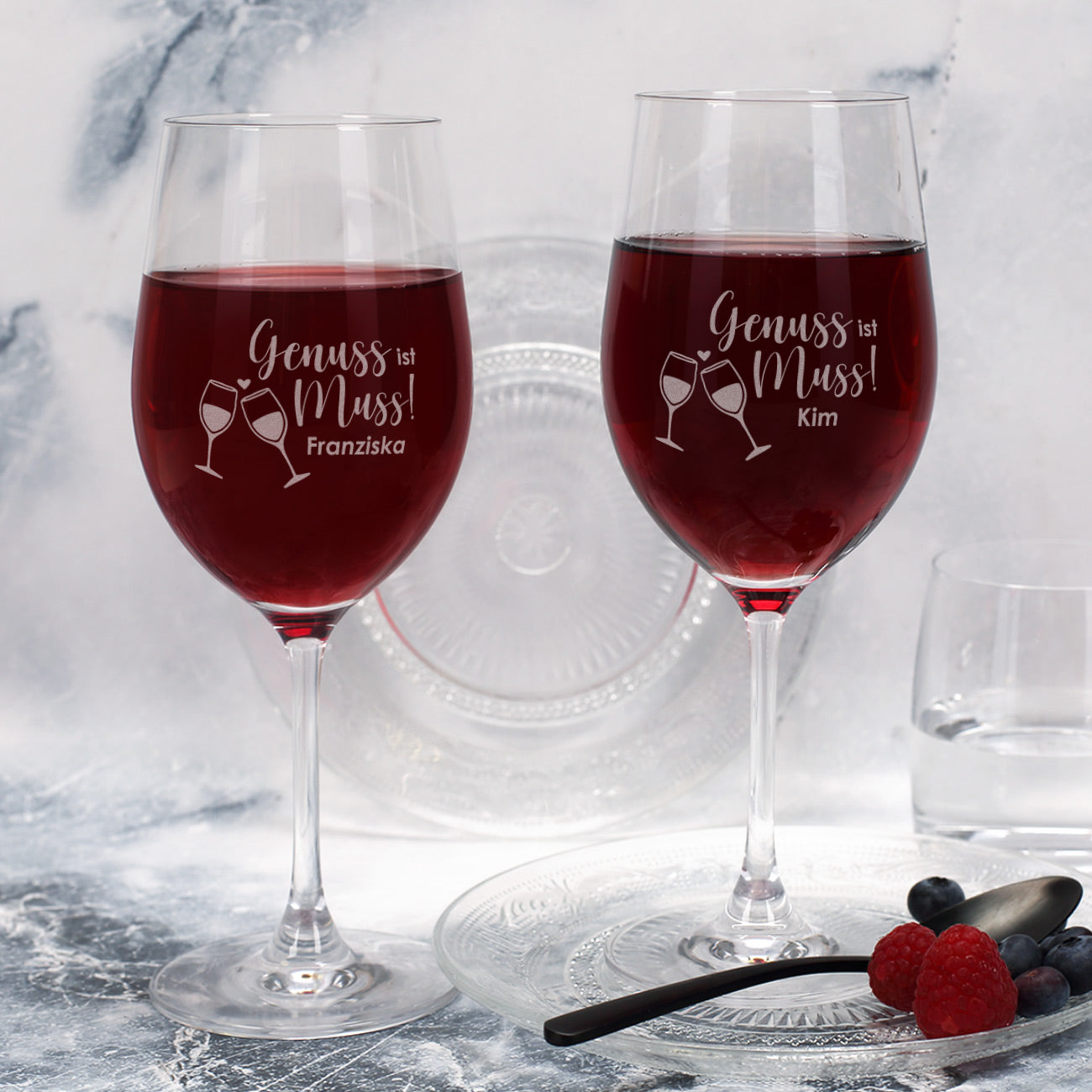 Zwei gravierte Weingläser „Genuss ist Muss“ mit Namen, befüllt mit Rotwein, auf Tisch mit Teller und Beeren