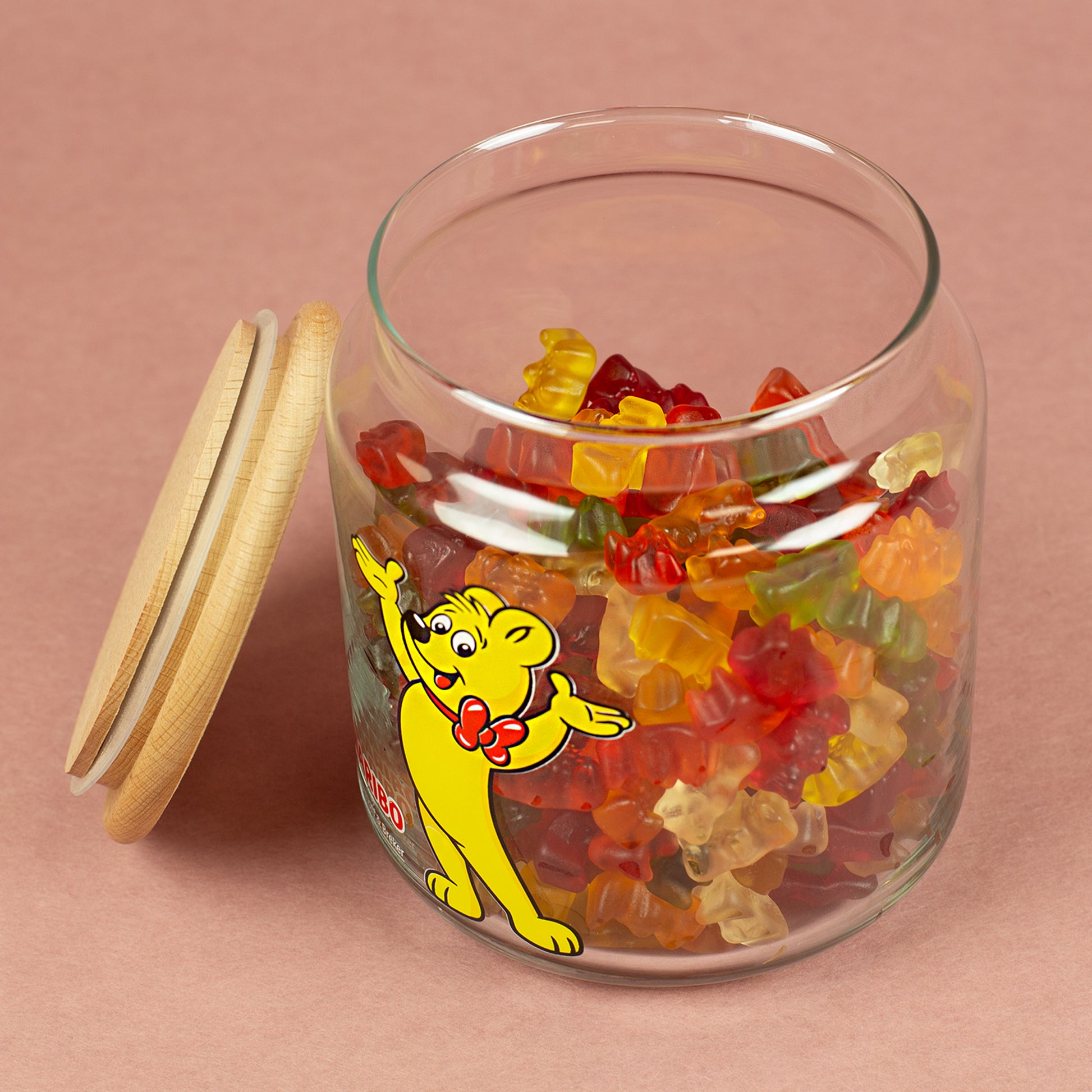 Offene HARIBO Glasdose mit Goldbär-Motiv, gefüllt mit bunten Gummibärchen, Holzdeckel seitlich liegend.