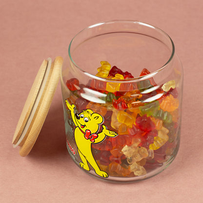 Offene HARIBO Glasdose mit Goldbär-Motiv, gefüllt mit bunten Gummibärchen, Holzdeckel seitlich liegend.