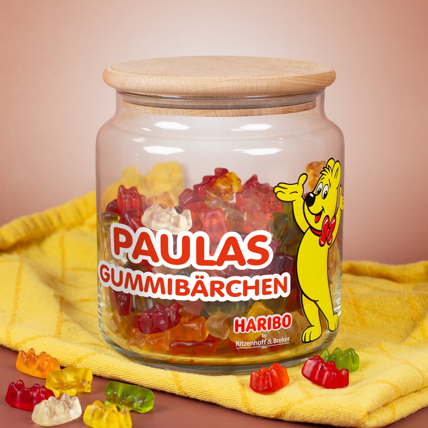 Personalisierbares HARIBO Vorratsglas gefüllt mit bunten Gummibärchen, Name ‚Paulas Gummibärchen‘ im roten Druck, Goldbär-Motiv, Holzdeckel daneben auf gelbem Tuch.