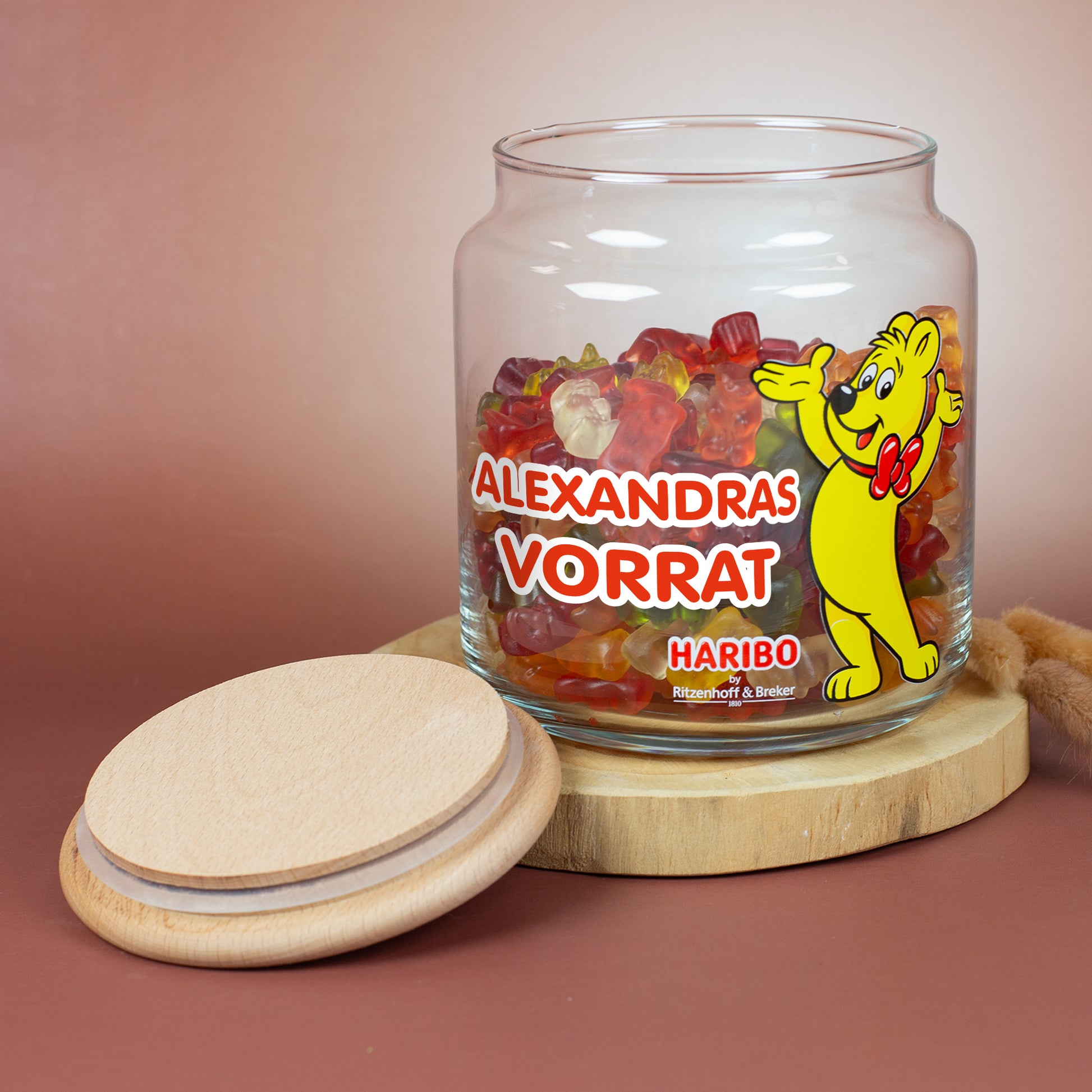 HARIBO Vorratsglas mit Goldbär und personalisiertem Text ‚Alexandras Vorrat‘, gefüllt mit Gummibärchen, Holzdeckel daneben auf rundem Holzbrett.
