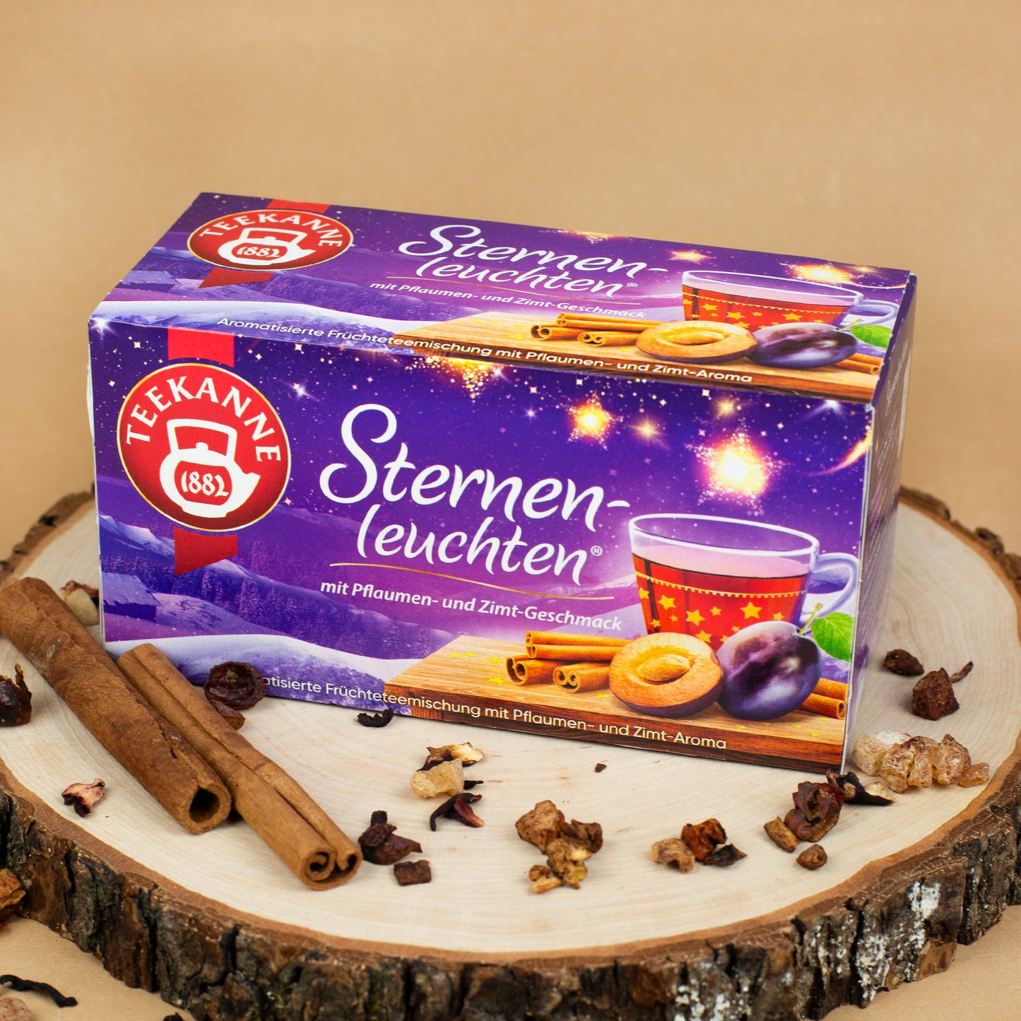 Packung „Teekanne Sternenleuchten“ in violettem Nachthimmel-Design mit Pflaume, Gebäck und Zimtstangen, dekorativ arrangiert auf einer Holzscheibe.