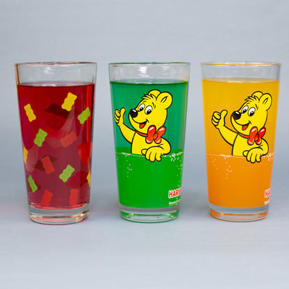 Drei HARIBO Trinkgläser mit bunten Motiven und farbigen Getränken als Vergleichsansicht auf grauem Hintergrund