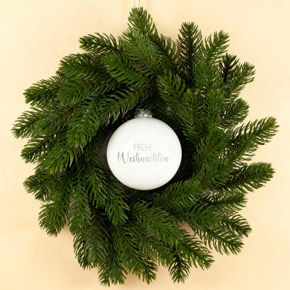 Weiß glänzende Weihnachtskugel mit Gravur Frohe Weihnachten im Türkranz auf hellem Hintergrund