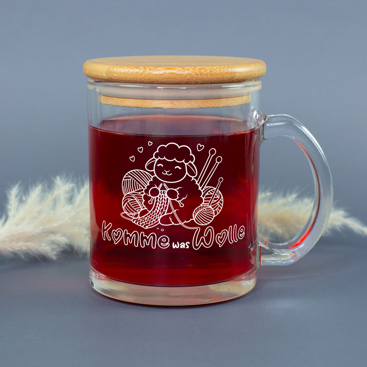 Glastasse mit rotem Tee, Bambusdeckel und weißem Strickmotiv mit Schaf, Wolle und Stricknadeln