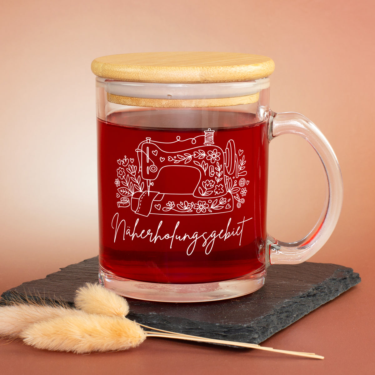 Glastasse mit rotem Tee, Bambusdeckel und weißem Nähmaschinen-Motiv „Näherholungsgebiet“ auf Schieferplatte