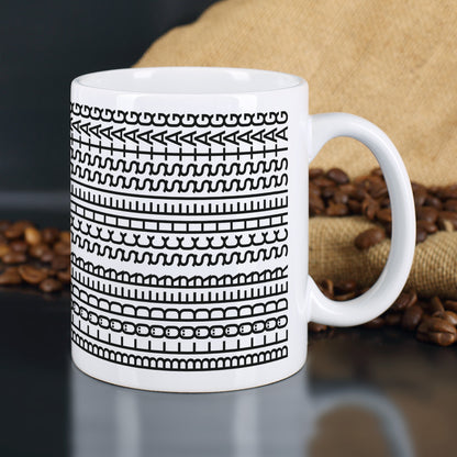 Weiße Keramiktasse mit schwarzem Muster und verstecktem Spruch vor Kaffeesack und Kaffeebohnen