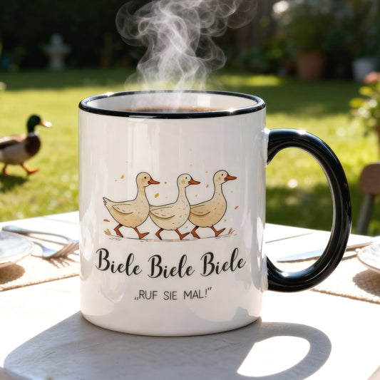 Tasse mit Entenmotiv und Spruch „Biele Biele Biele – Ruf sie mal!“ auf Gartentisch, im Hintergrund grüne Wiese mit Ente.