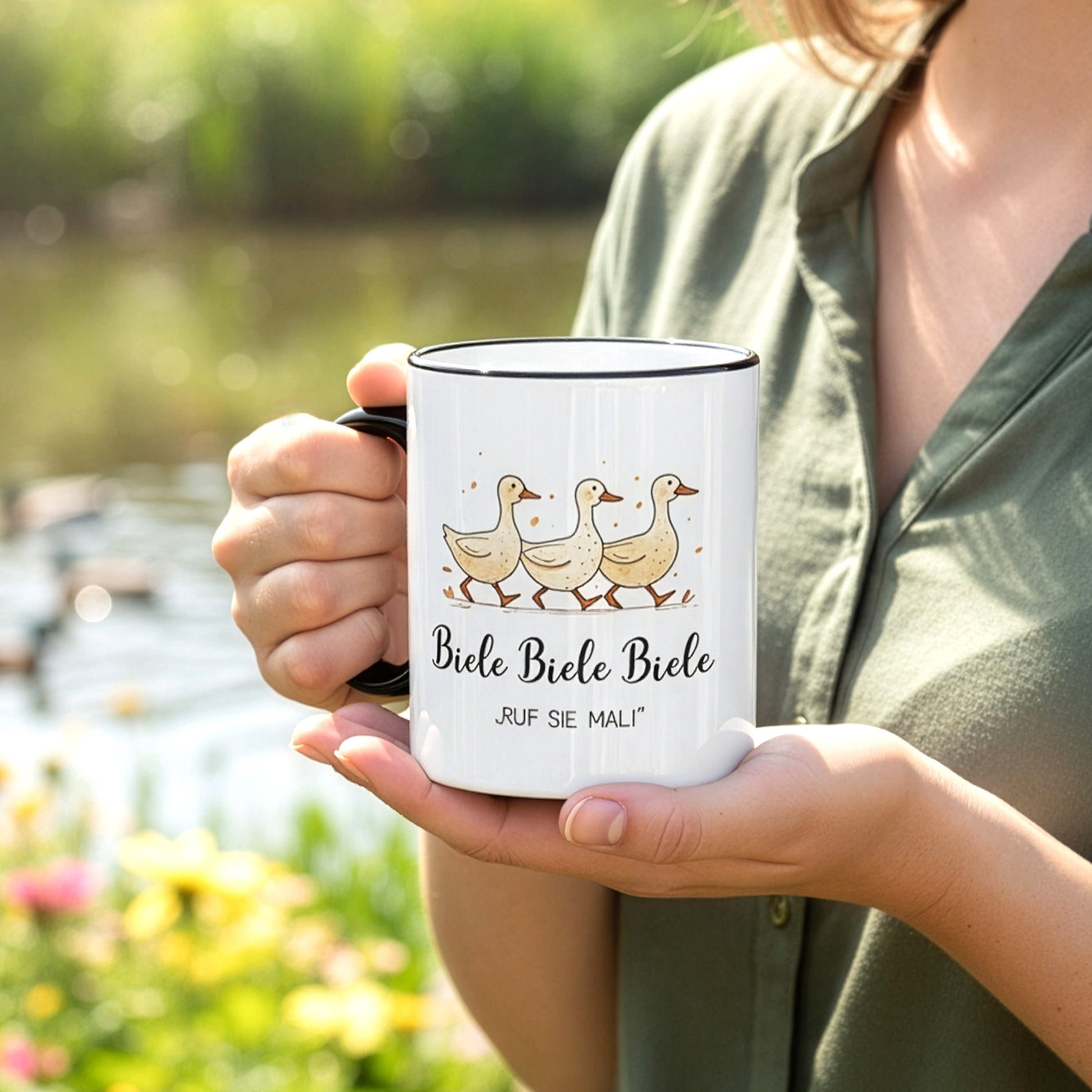 Frau hält Tasse mit Entenmotiv und Spruch „Biele Biele Biele – Ruf sie mal!“ vor Teich mit Enten im Hintergrund.