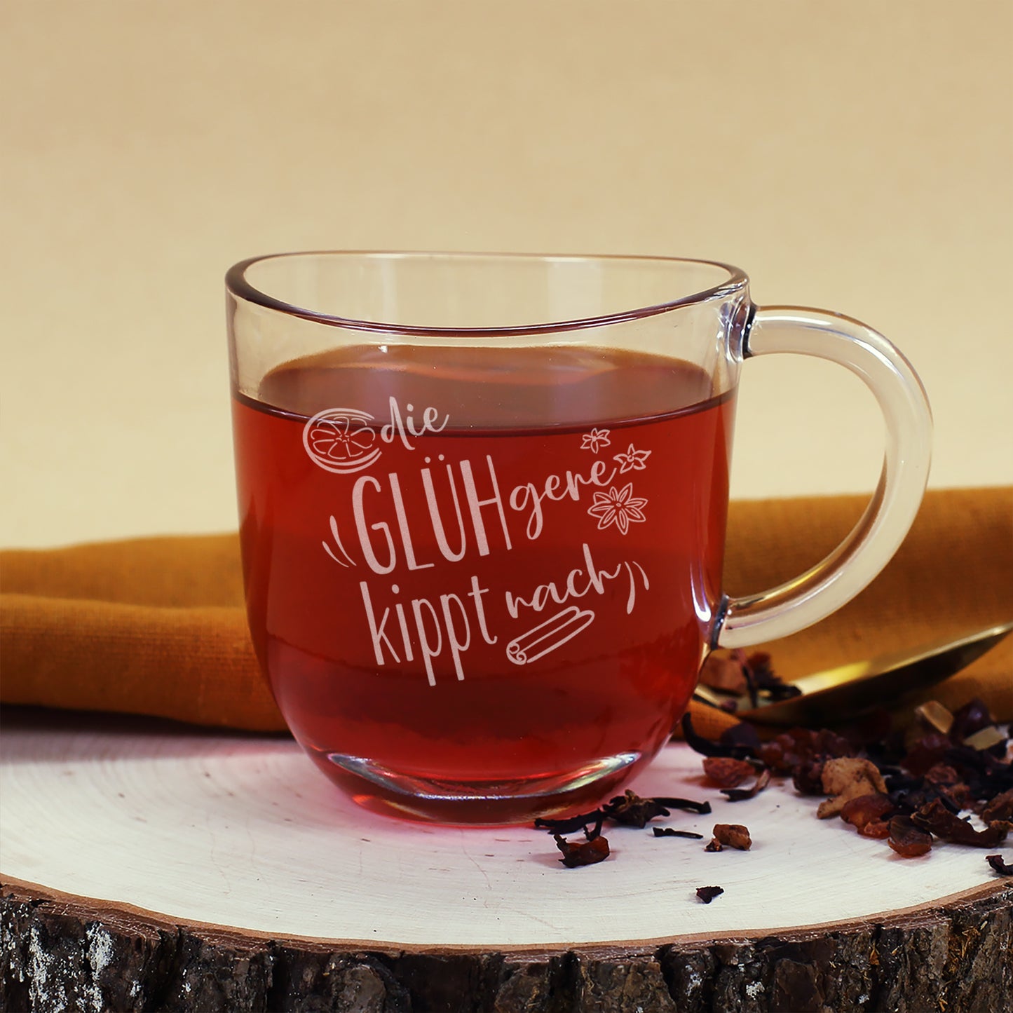 Leonardo Glühweintasse „Die GLÜHgere kippt nach“, befüllt mit Glühwein, auf Holzscheibe mit goldfarbenem Löffel und Teezutaten dekoriert