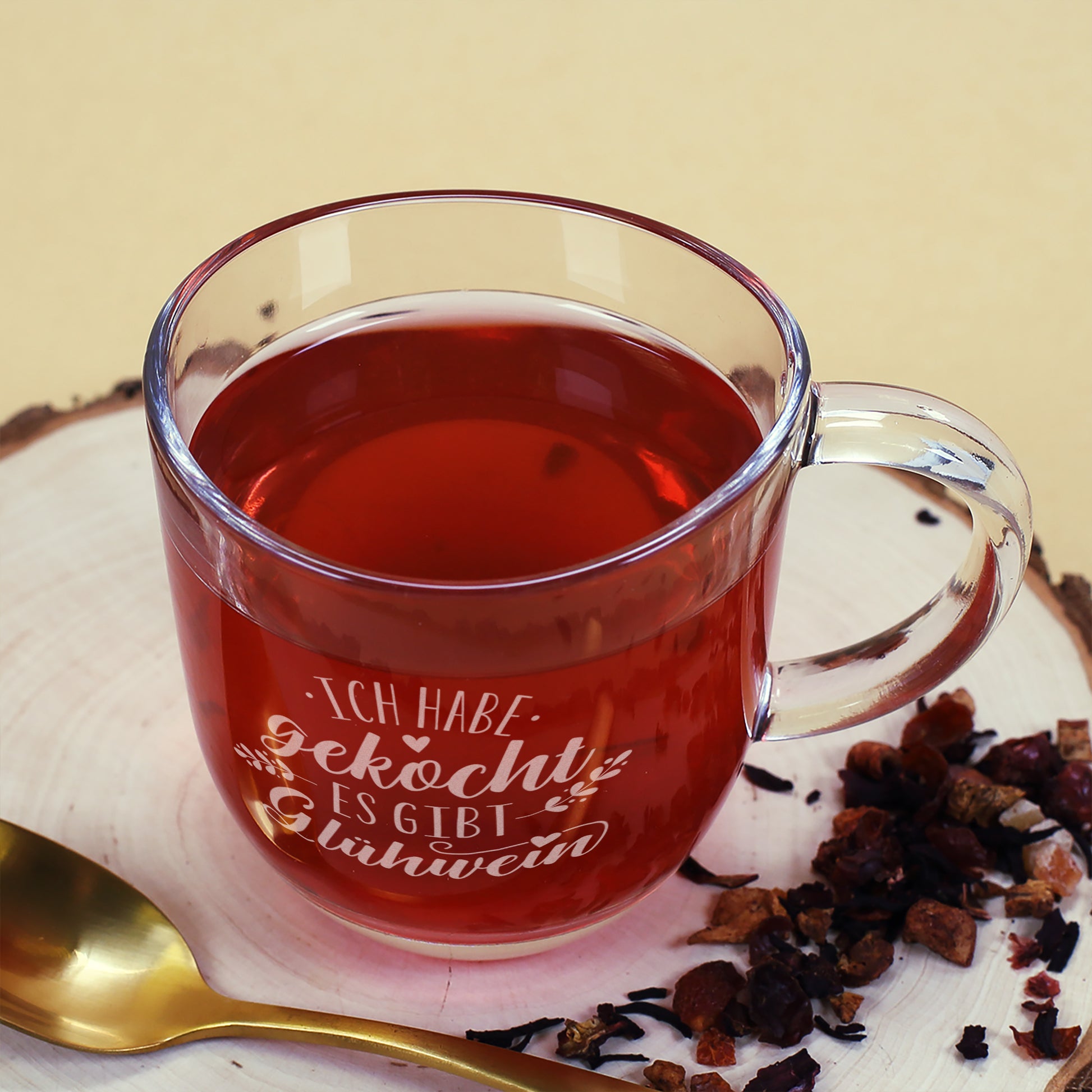 Glühweintasse aus Glas mit Gravur, gefüllt mit rotem Getränk, auf hellem Holzbrett mit goldfarbenem Löffel und winterlichen Teezutaten