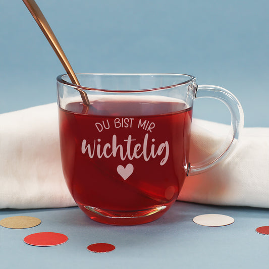Seitlich fotografiertes Teeglas mit Gravur „Du bist mir wichtelig“, gefüllt mit rotem Tee vor blauem Hintergrund.