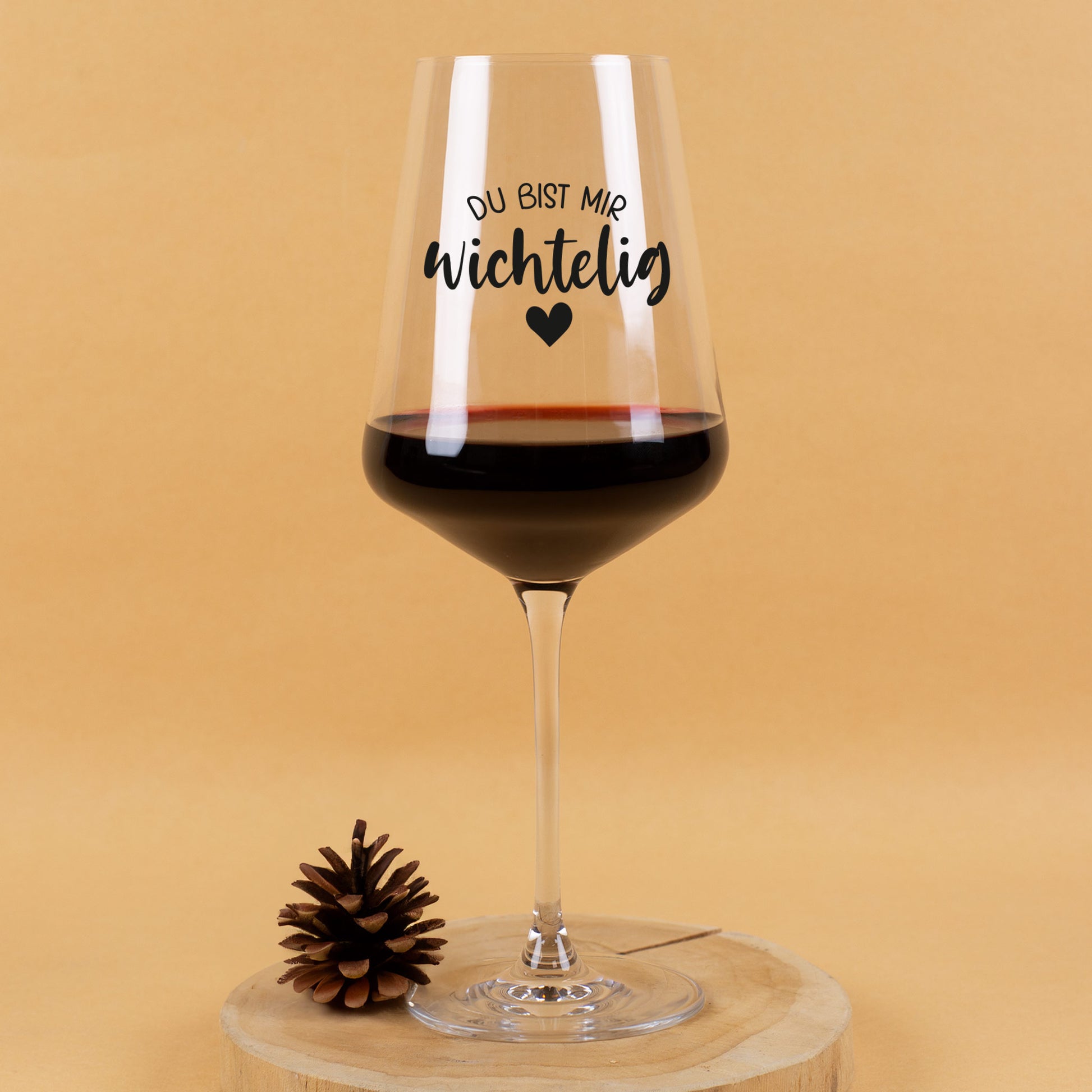 Leonardo Weinglas mit schwarzem Aufdruck „Du bist mir wichtelig“ und Herzmotiv, gefüllt mit Rotwein, dekoriert mit Tannenzapfen