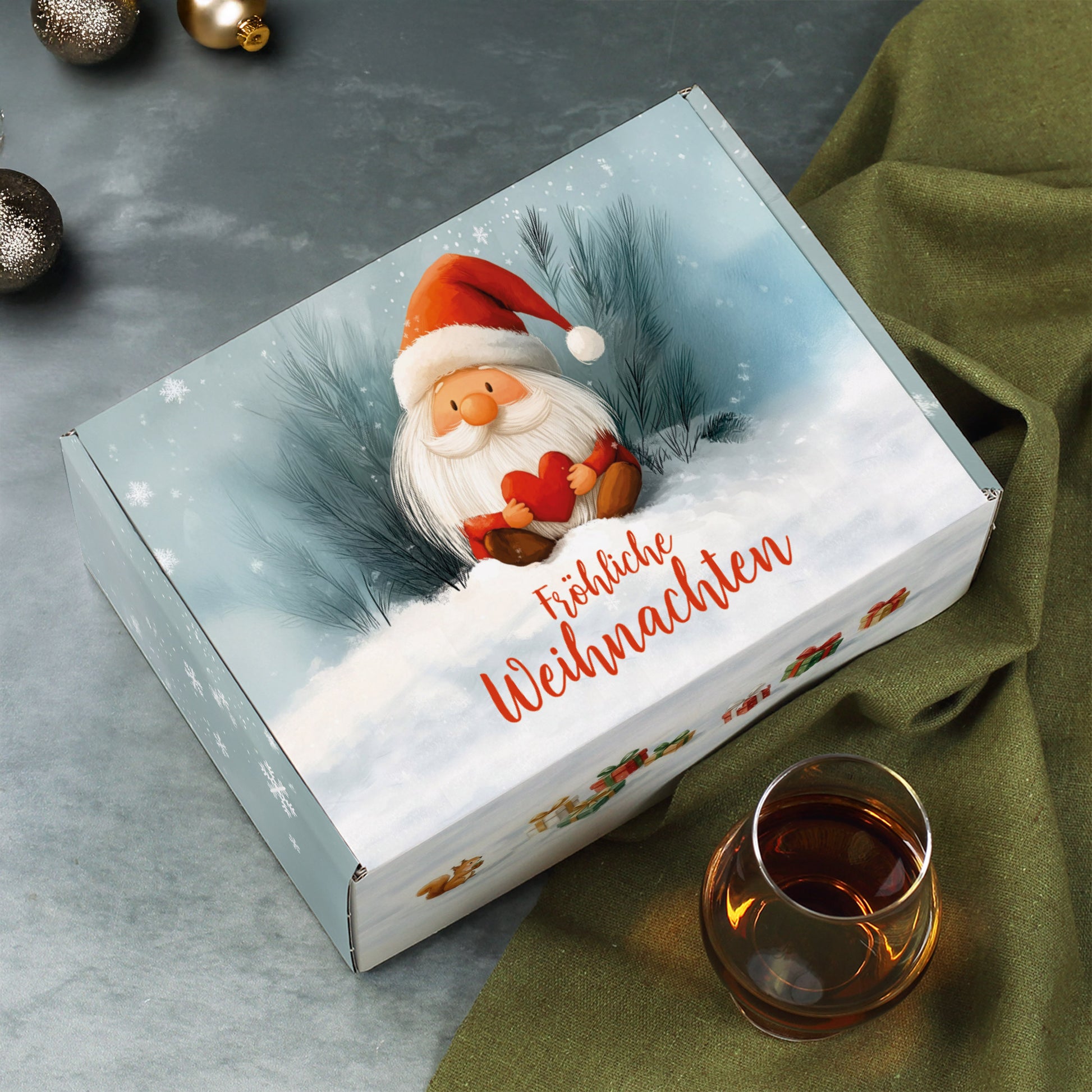Weihnachts-Geschenkbox mit Wichtel im Schnee – festlich arrangiert neben grüner Stoffdecke und Glas