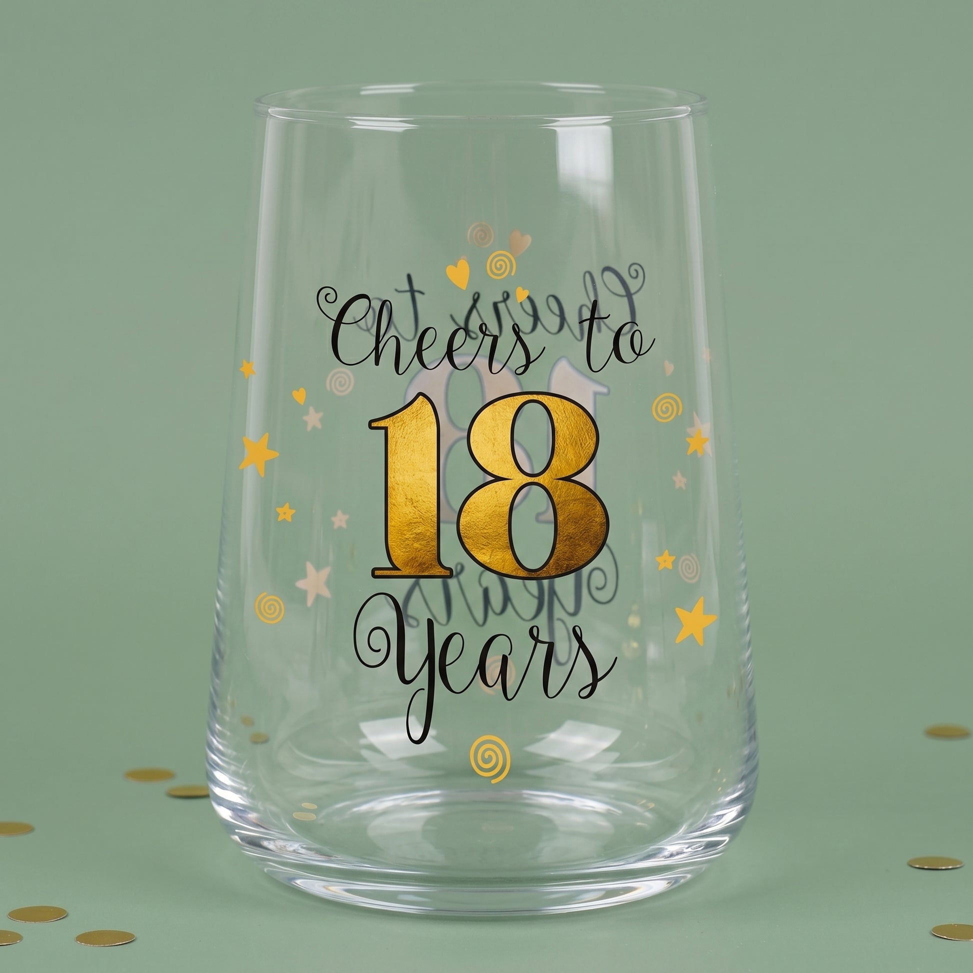 Leeres Trinkglas zum 18. Geburtstag mit goldener 18, Sternen und Konfetti auf grünem Hintergrund