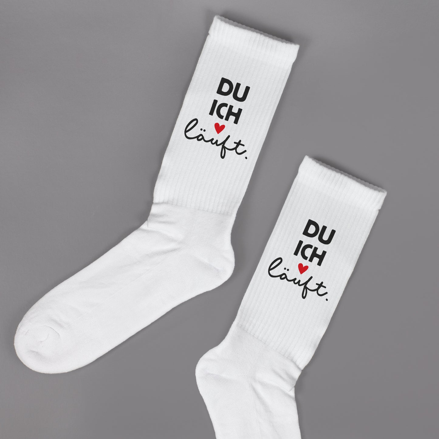 Zwei weiße Socken liegend auf grauem Hintergrund, Aufdruck „DU ICH läuft.“ mit Herz gut sichtbar