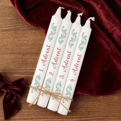 Vier weiße Adventskerzen mit rotem Schriftzug 1.–4. Advent und grün-rotem Zweigmotiv, dekorativ auf Holz und rotem Stoff arrangiert.