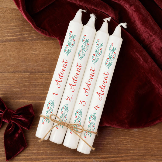 Vier weiße Adventskerzen mit rotem Schriftzug 1.–4. Advent und grün-rotem Zweigmotiv, dekorativ auf Holz und rotem Stoff arrangiert.