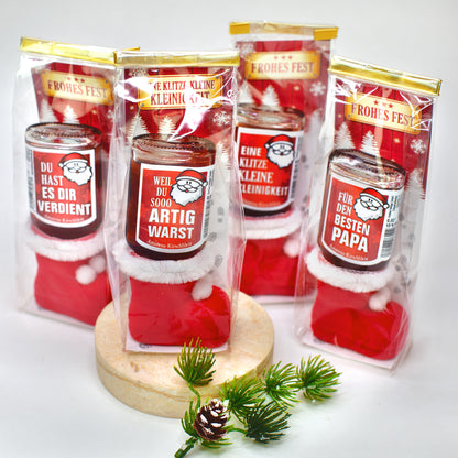 Vier rote Nikolausstiefel mit je einer auf dem Kopf stehenden Likörflasche, verpackt in Klarsichtbeuteln mit Goldverschluss. Jeder Likör trägt ein anderes Etikett mit Sprüchen wie „Artig warst“ oder „Bester Papa“. Im Hintergrund Weihnachtsmotive und Dekozweig.