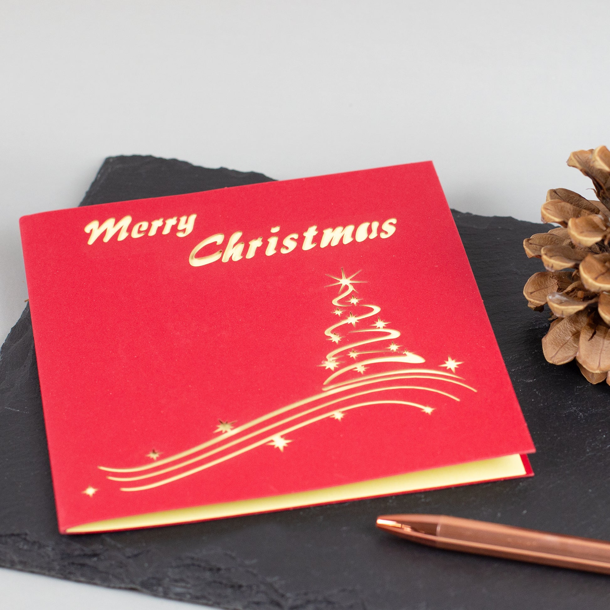 Geschlossene rote Weihnachtskarte mit stilisiertem Tannenbaum aus goldenen Linien und Schriftzug „Merry Christmas“.