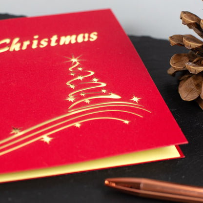 Detailaufnahme der Vorderseite der roten Weihnachtskarte mit goldenem Tannenbaum-Design und „Merry Christmas“-Schriftzug.