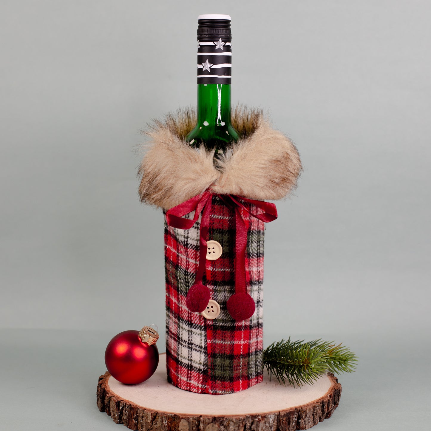 Flaschen Geschenkverpackung in Mantel-Optik, kariertes Muster in Rot-Grün, mit Pelzkragen und roten Schleifen, auf Holzplatte mit Tannenzweig.