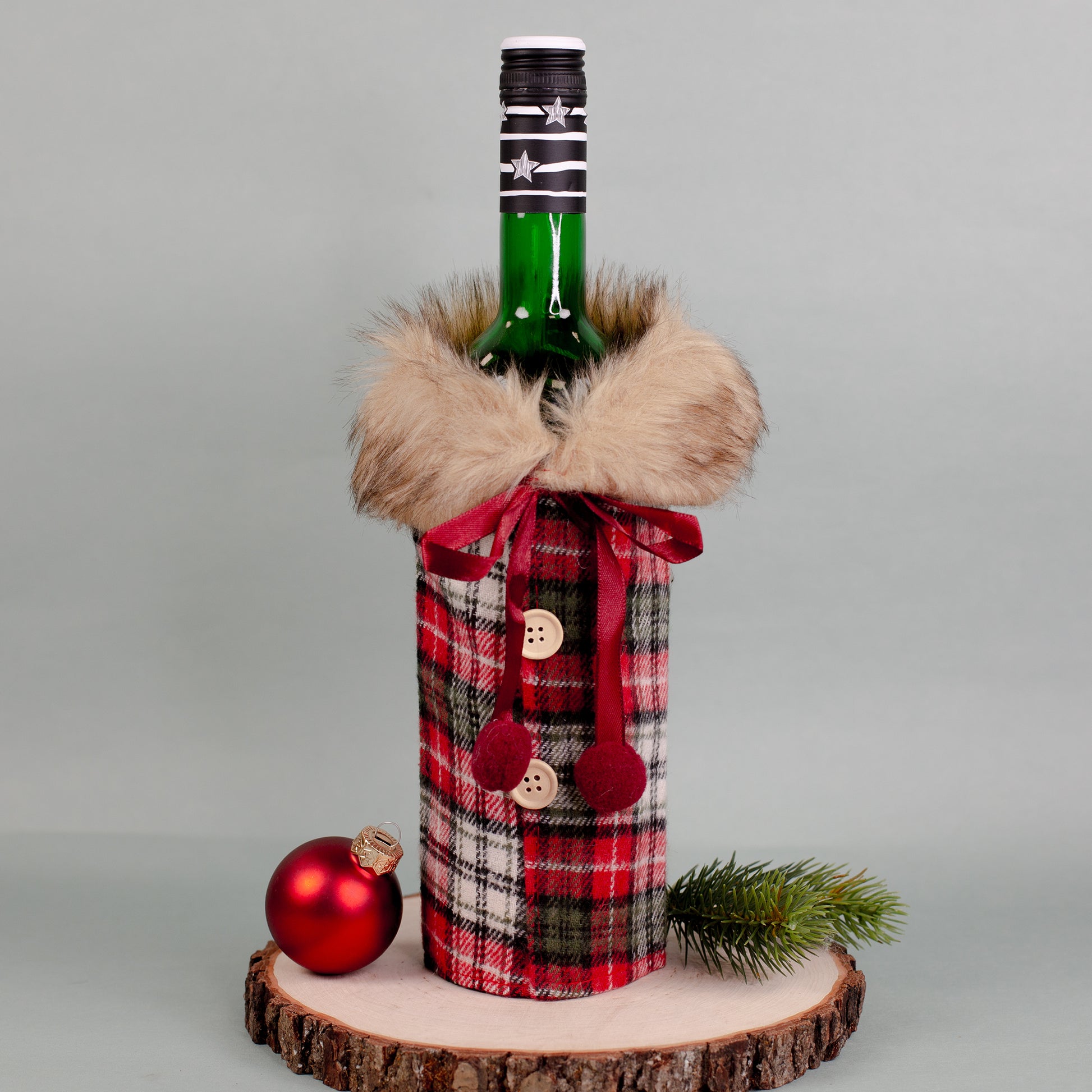 Flaschen Geschenkverpackung in Mantel-Optik, kariertes Muster in Rot-Grün, mit Pelzkragen und roten Schleifen, auf Holzplatte mit Tannenzweig.