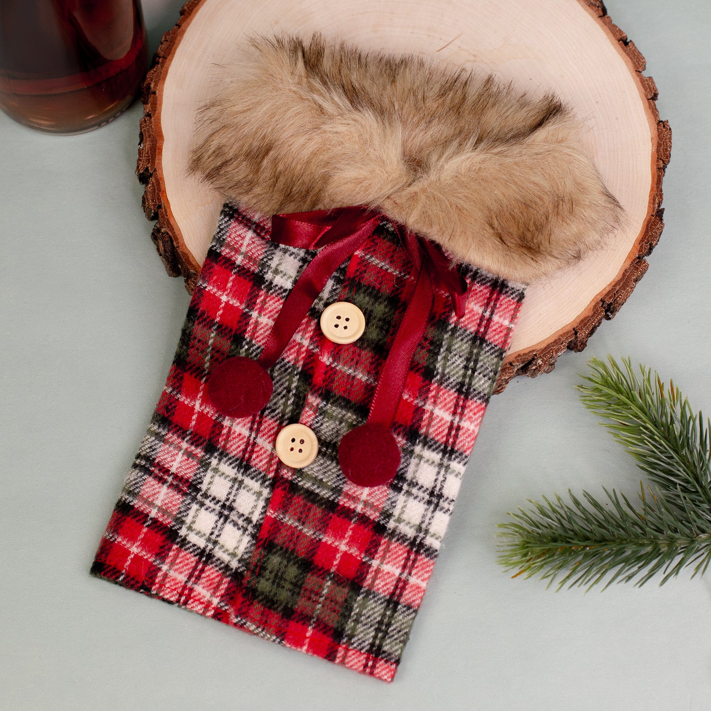 Kariertes Flaschenmäntelchen mit Pelzkragen flach auf Holzscheibe liegend, neben Flasche.