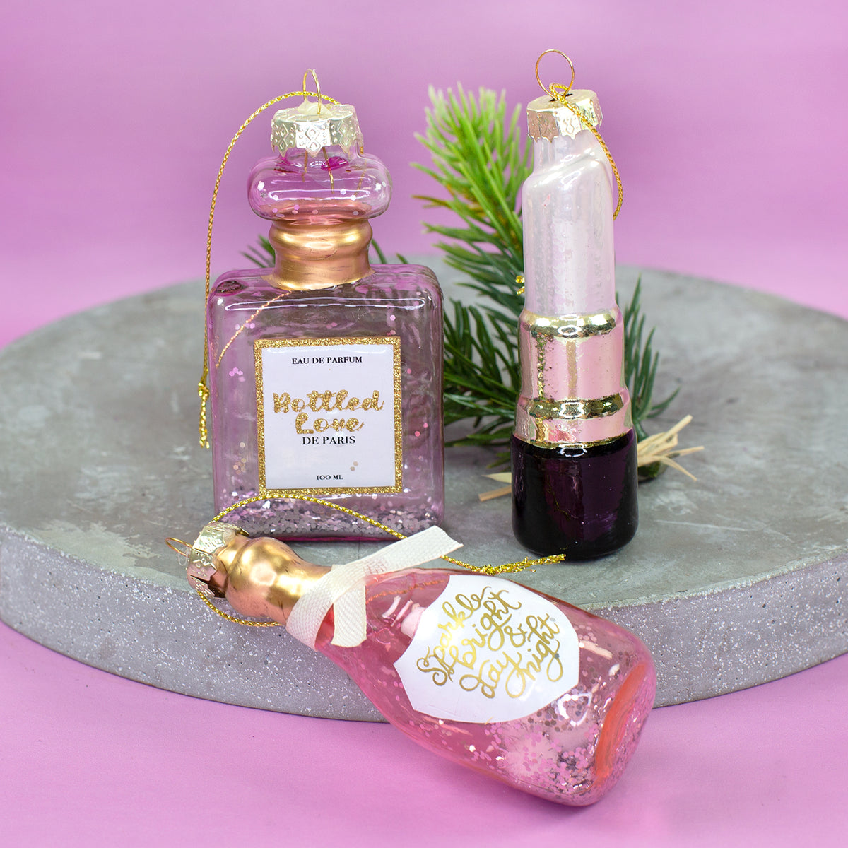 Drei rosa-goldene Glas Weihnachtsanhänger mit Parfümflakon, Lippenstift und Sektflasche dekoriert