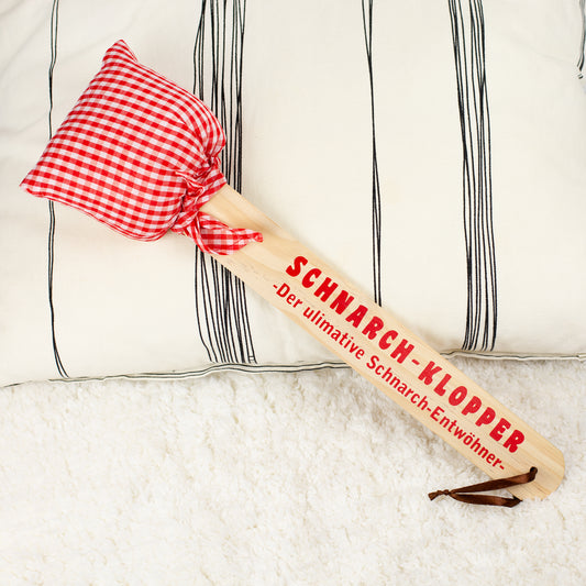 Schnarch-Klopper mit Holzgriff und rot-weiß kariertem Stoffkissen, Aufdruck „Der ultimative Schnarch-Entwöhner“, liegend auf hellem Untergrund