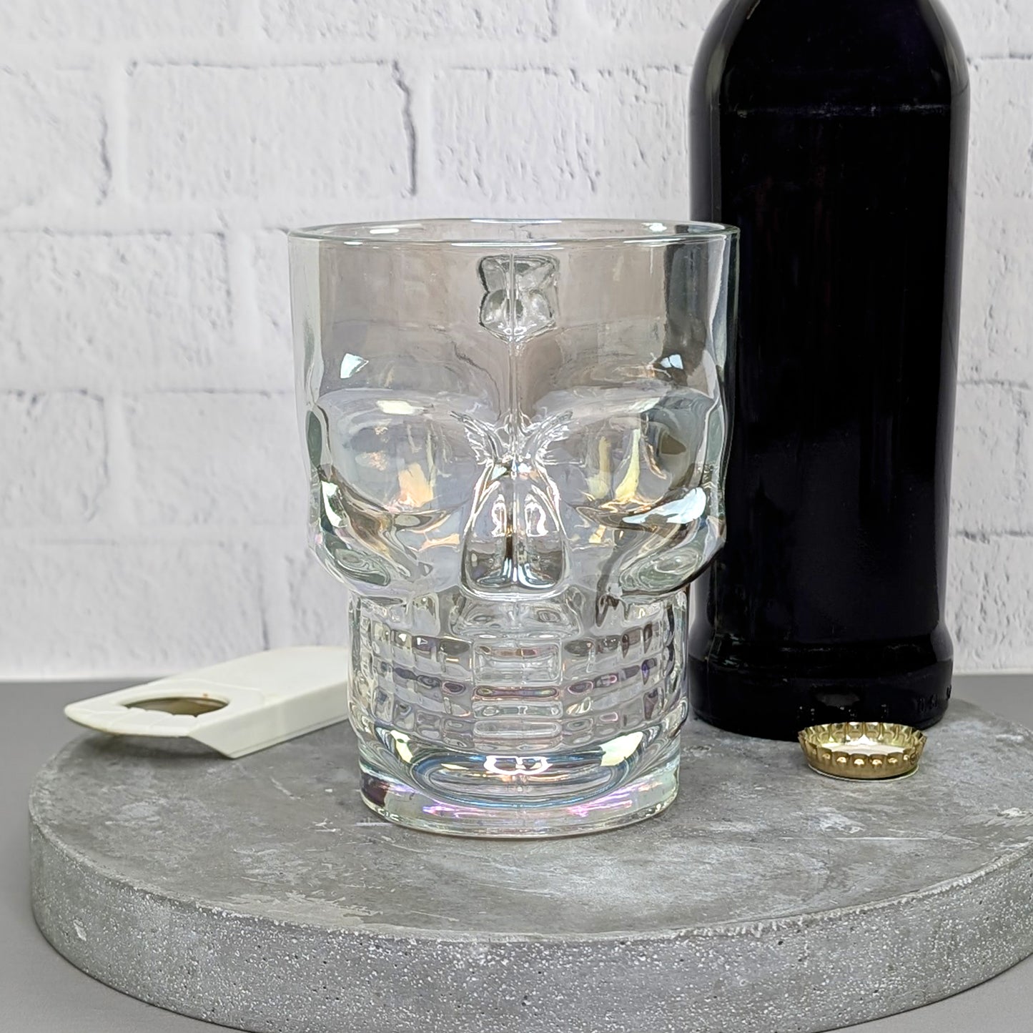 Totenkopfglas frontal, leer auf Betonplatte mit Bierflasche im Hintergrund, Öffner und Kronkorken