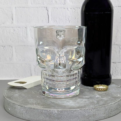 Totenkopfglas frontal, leer auf Betonplatte mit Bierflasche im Hintergrund, Öffner und Kronkorken