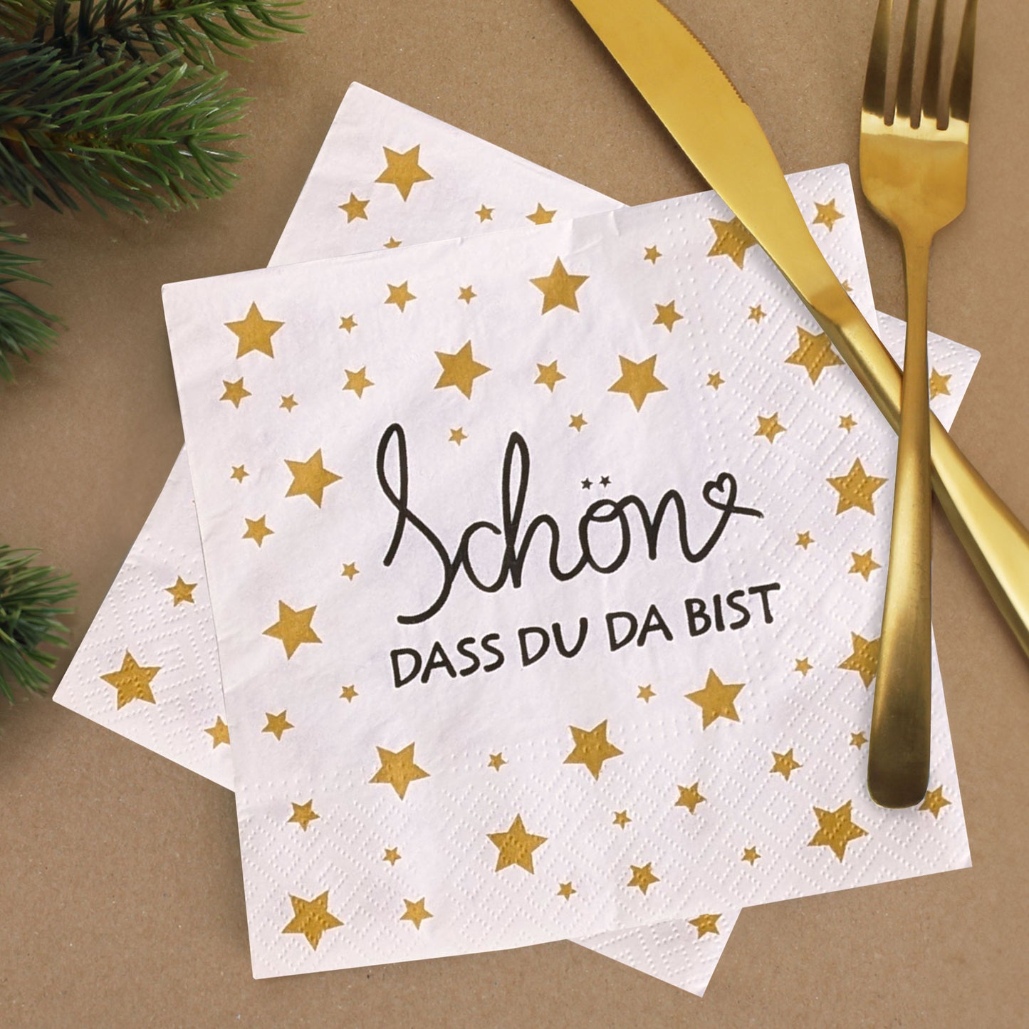 Weiße Servietten mit goldenem Sternenmuster und Schriftzug ‚Schön, dass du da bist‘, festlich mit goldfarbenem Besteck und Tannenzweigen dekoriert.