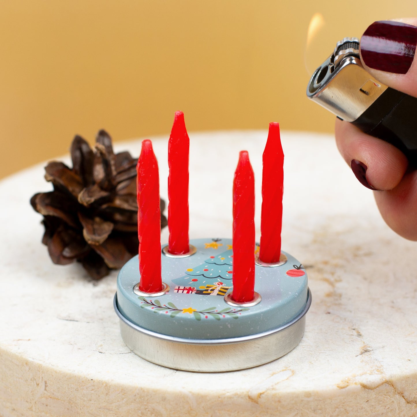 Eine Hand mit Feuerzeug zündet eine der vier roten Kerzen auf einem Mini-Adventskranz mit Weihnachtsbaum-Motiv an, dekoriert mit Tannenzapfen.