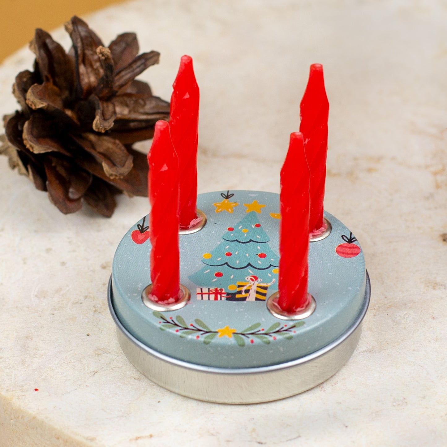 Mini-Adventskranz mit Weihnachtsbaum-Motiv und vier eingesetzten roten Kerzen, auf einer Steinoberfläche mit Tannenzapfen im Hintergrund.